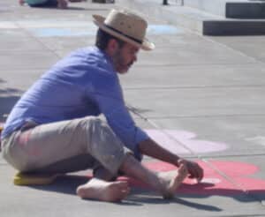 man in straw hat blue shirt tan pants sitting on pavement drawing at sidewalk chalk festival