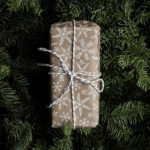 Rectangular shaped gift wrapped in beige and white snowflake paper tied with bow of white string, evergreens behind