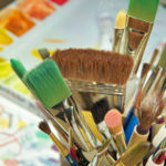 Closeup view from above of cluster of different size and color brushes – pink, green, brown, yellow – above artwork