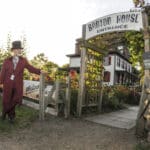 man in top hat & 1850s long burgundy coat standing by gateway to Barton House in background