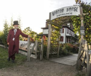 man in top hat & 1850s long burgundy coat standing by gateway to Barton House in background