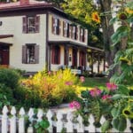 white picket fence and blooming flowers including sunflowers in front of Barton House with cream paint and burgundy shutters