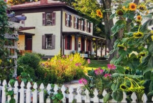 white picket fence and blooming flowers including sunflowers in front of Barton House with cream paint and burgundy shutters