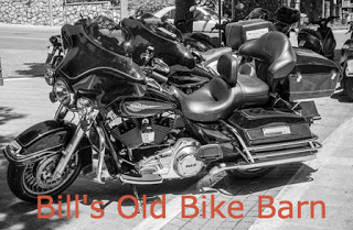 black and white motorcycles in a row on gray pavement Orange text: Bill's Old Bike Barn