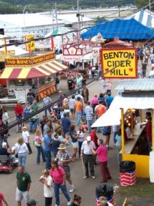 Birds-eye view of county fair with crowd walking between two rows of red/yellow, red/white booths, blue, blue/white striped tents