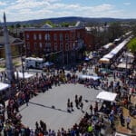 Overhead view of gray town square surrounded by people on all four sides, street with white tents down middle