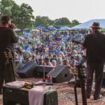 musicians on outdoor stage with backs to camera playing to large crowd with green trees and white tents behind