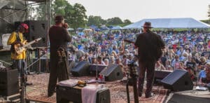 musicians on outdoor stage with backs to camera playing to large crowd with green trees and white tents behind