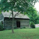 Green lawn with gray and white log Quaker meetinghouse built 1789 large tree shading it