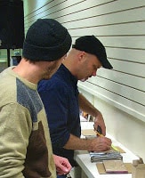 Man in black knit cap and multicolor brown and gray sweater watching man in blue shirt and black ballcap measure materials on ledge in front of white wall