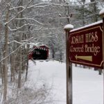 Burgundy and gold sign for Josiah hes Covered Bridge in foreground, snow covered trees and red bridge in background