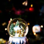 snowglobe of snow covered evergreen trees, cottage, snowman, with back hat children at night with holiday tree lights behind