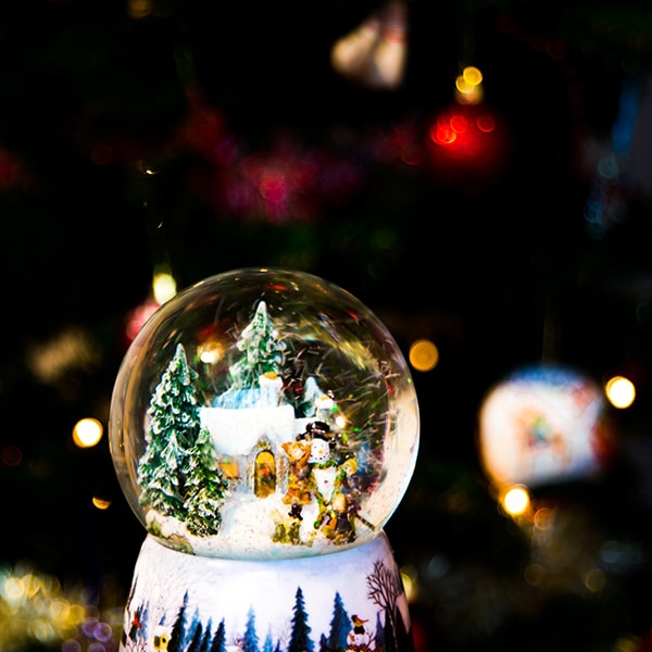 snowglobe of snow covered evergreen trees, cottage, snowman, with back hat children at night with holiday tree lights behind