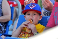 Danville young boy in blue Crosscutters cap and yellow shirt eating hot dog  held in both hands, crowd behind!