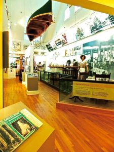 two story museum, canoe suspended from ceiling, pictures along white walls, restaurant exhibit with waitress behind waist high smoky glass