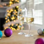2 glasses white wine and Christmas balls on table with lighted Christmas tree and window with blinds behind