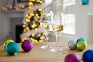 2 glasses white wine and Christmas balls on table with lighted Christmas tree and window with blinds behind