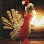 Sculpture of turkey wearing red bow around neck and Santa hat, standing in front of blazing fireplace