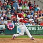 baseball player in white uniform with red sleeves and helmut hitting baseball with fans in stands behind