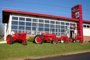 Three red tractors & International Harvester prototype dealership with façade of large multi-paned glass and red roof and tower displaying IH logo