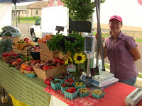 woman in red ballcap, shirt standing behind long table covered with farm fruits, vegetables, scale, green check and red tablecloths