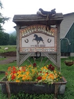 Sign for Brace's Stables with silhouette of horse and 2 horseshoes in center, shingle roof at top, red, orange flowers and green foliage in trough at bottom of sign