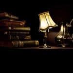 stack of old books, lighted antique lamp with tilted cream shade, silver pitcher on table in dark room