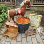 Antique brown rocking horse with cream mane and tail, red saddle, wood bucket, framed sampler, wooden boxes on brick sidewalk