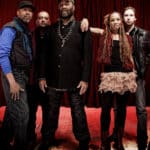 Four men and a woman with long curly hair dressed mostly in black standing in front of red curtain