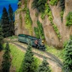 Model train on tracks next to mountainside with evergreens and gray rock cliffs