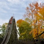 People riding cars on wooden roller coaster surrounded by orange and red trees during fall foliage season
