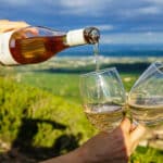 hand holding wine bottle pouring wine into two glasses against background landscape