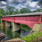 red covered bridge over quiet river. Gay roof and limestone piers, green trees and cloudy sky behind, portion of ties of railroad bridge in foreground