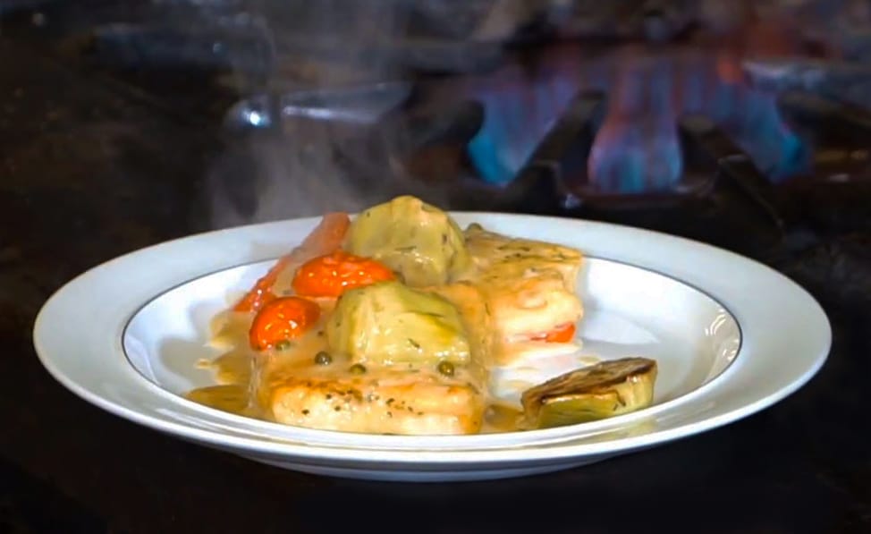 Chilean Sea Bass with artichokes and tomatoes cream sauce on white plate in front of gas flame on black stove