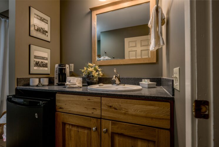 Photo shows bathroom vanity which includes small refrigerator, coffee maker, large mirror and two photos on the wall.