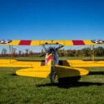 Vintage Steerman yellow bi-plane with red & yellow striped wings on grassy airfield