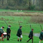 5 soldiers dressed in Revolutionary War uniforms with rifles on shoulders marching in a line on green grass