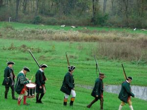 5 soldiers dressed in Revolutionary War uniforms with rifles on shoulders marching in a line on green grass