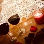 diagonal of two glasses of red wine, chocolates, burning candle in red holder, rose petal on wood table with white geometric background