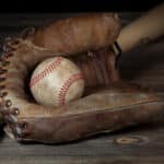 Dirty baseball nested in brown baseball glove with bat at angle behind on wooden surface