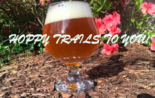 footed glass of beer with creamy head on brown mulch ground, pink flowers and green foliage behind Text: Happy Trails to You!