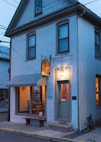Two story bluish gray building with dark trim at dusk, yellow light through picture window with awning, sign and light over doorway, three windows above
