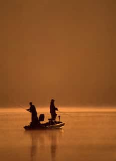 sepia silhouette of two fishermen standing in small boat on still lake with dark sky behind