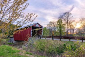 Kramer Covered Bridge in summer - red bridge with brown wood guardrails along road leading to bridge - trees surrounding and blue sky with clouds