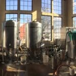 interior of brewery with stainless tanks, man on ladder by tank and large windows showing blue sky