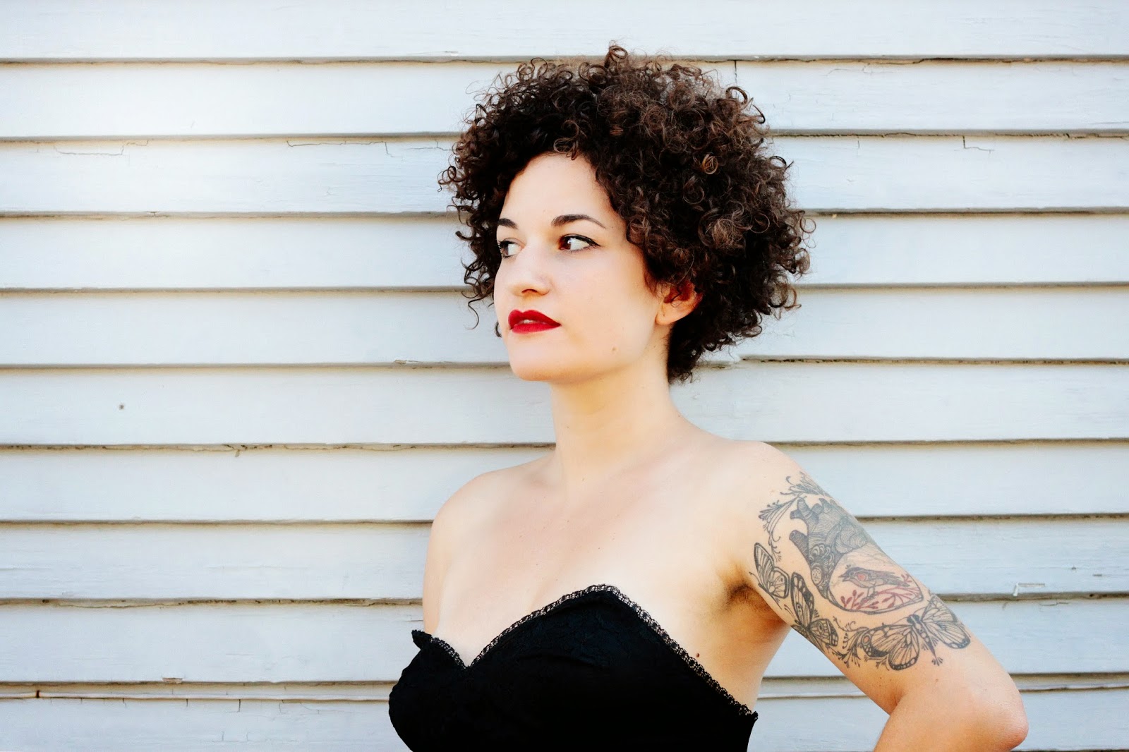 Closeup of head and torso of woman with black curly hair, red lipstick, tattoos on left arm, strapless black dress