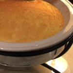 Closeup of golden cheesy grits in cream color round casserole with black wrought iron holder