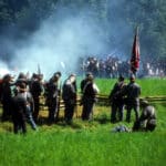 Civil War re-enactors standing behind wood rail fence firing volley with white smoke rising over green field