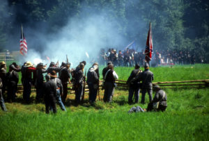 Civil War re-enactors standing behind wood rail fence firing volley with white smoke rising over green field