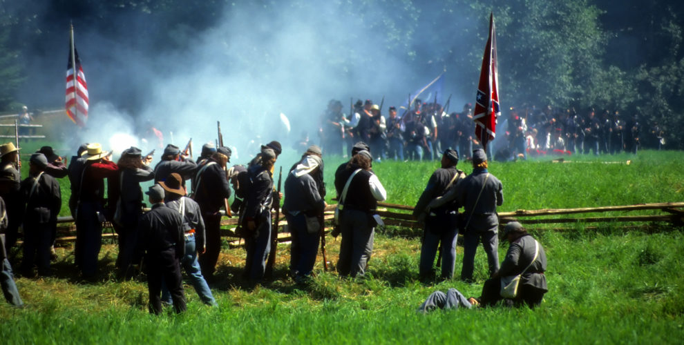 Civil War re-enactors standing behind wood rail fence firing volley with white smoke rising over green field